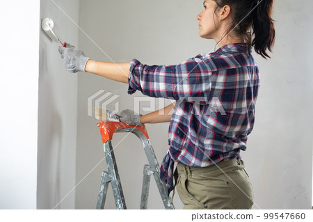 Woman in paint roller and white paint paints the wall of house on stepladder. Construction work and cosmetic repairs in house, wall painting, tinting, finishing work with your own hands Woman in paint roller and white paint paints the wall of house on stepladder. Construction work and cosmetic repairs in house, wall painting, tinting, finishing work with your own hands 99547660