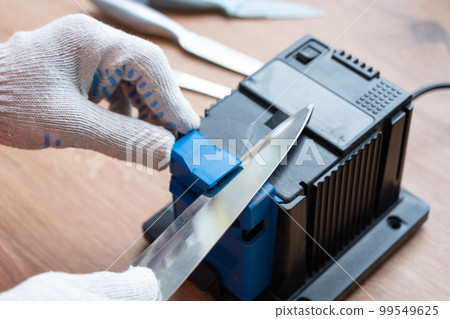 Sharpening a knife on an electric sharpener at home. The man's hand drives the knife blade between the blue sharpeners, dust flies on the machine. Sharpening a knife on an electric sharpener at home. The man's hand drives the knife blade between the blue sharpeners, dust flies on the machine. 99549625