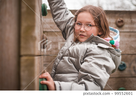 Primary school girl is climbing a rock wall at the playground. Sport time at school. Child in glasses. Save and protect. Kid outdoors Primary school girl is climbing a rock wall at the playground. Sport time at school. Child in glasses. Save and protect. Kid outdoors 99552601