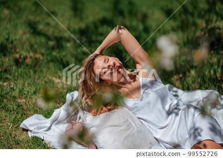 woman sleeps on a white bed in the fresh spring grass in the garden. Dressed in a blue nightgown. woman sleeps on a white bed in the fresh spring grass in the garden. Dressed in a blue nightgown. 99552726