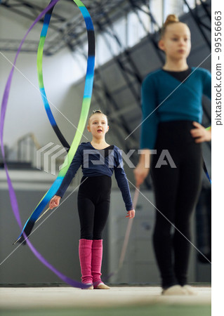 Little girls, children, rhythmic gymnasts training indoors, doing exercise with ribbon. Concept of sport, childhood, rhythmic gymnastics Little girls, children, rhythmic gymnasts training indoors, doing exercise with ribbon. Concept of sport, childhood, rhythmic gymnastics 99556663