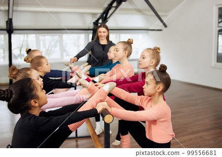 Group of little girls, children, gymnasts training with coach indoors. Stretching exercises with barre equipment. Concept of sport, childhood, rhythmic gymnastics Group of little girls, children, gymnasts training with coach indoors. Stretching exercises with barre equipment. Concept of sport, childhood, rhythmic gymnastics 99556681
