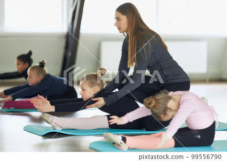 Woman stretching kids on matt. Flexibility. Group of little girls, children, gymnasts training with coach indoors. Concept of sport, childhood, rhythmic gymnastics Woman stretching kids on matt. Flexibility. Group of little girls, children, gymnasts training with coach indoors. Concept of sport, childhood, rhythmic gymnastics 99556790