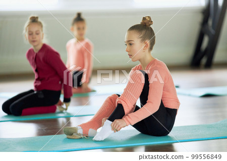 Little girls, children sitting on matt and preparing for sports training session indoors. Warming-up exercises. Concept of sport, childhood, rhythmic gymnastics Little girls, children sitting on matt and preparing for sports training session indoors. Warming-up exercises. Concept of sport, childhood, rhythmic gymnastics 99556849