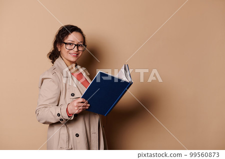 Charming middle-aged multi-ethnic woman wearing stylish spectacles and elegant beige coat, reads a book, smiles looking at camera, isolated on beige background with copy ad space Charming middle-aged multi-ethnic woman wearing stylish spectacles and elegant beige coat, reads a book, smiles looking at camera, isolated on beige background with copy ad space 99560873