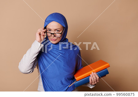 Strict Arabic Muslim beautiful middle-aged woman teacher educator in a blue hijab, looking at camera through her eyeglasses, standing isolated on beige background with a pile of books in her hands Strict Arabic Muslim beautiful middle-aged woman teacher educator in a blue hijab, looking at camera through her eyeglasses, standing isolated on beige background with a pile of books in her hands 99560875