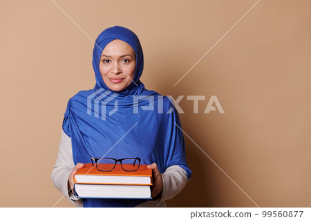 Charming Middle-Eastern Muslim woman - teacher, student, educator with head covered in blue hijab, holding pile of books and smiling confidently looking at camera, isolated on beige background Charming Middle-Eastern Muslim woman - teacher, student, educator with head covered in blue hijab, holding pile of books and smiling confidently looking at camera, isolated on beige background 99560877