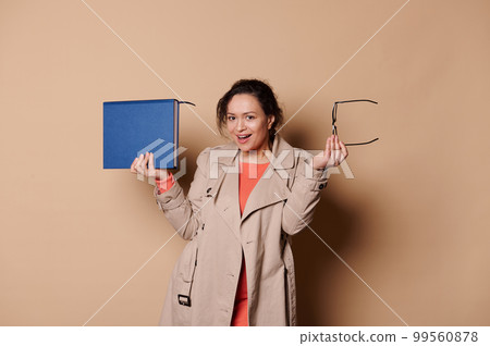 Happy positive middle-aged ethnic female teacher - educator - professor in elegant classical beige casual clothes, smiles and looks at camera while poses with a book and spectacles on cream background 99560878