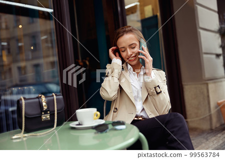 Smiling stylish woman sitting at cafe terrace talking on phone while drinking coffee Smiling stylish woman sitting at cafe terrace talking on phone while drinking coffee 99563784