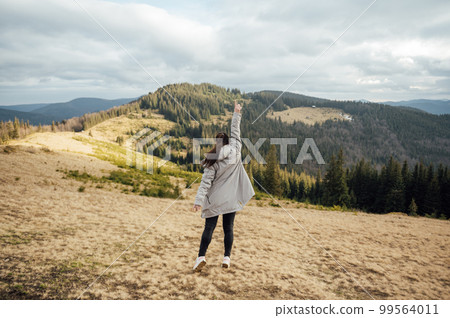 Happy girl stands in the mountains, enjoys the beautiful view and raises her hand up Happy girl stands in the mountains, enjoys the beautiful view and raises her hand up 99564011