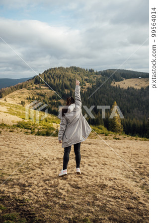 Happy girl stands in the mountains, enjoys the beautiful view and raises her hand up Happy girl stands in the mountains, enjoys the beautiful view and raises her hand up 99564014