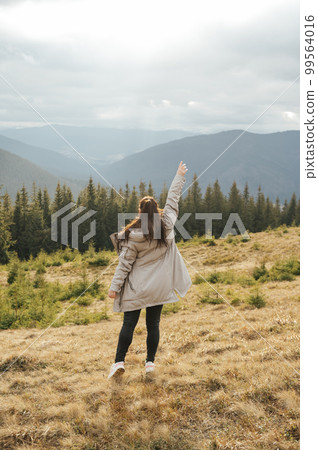 Happy girl stands in the mountains, enjoys the beautiful view and raises her hand up 99564016