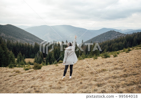 Happy girl stands in the mountains, enjoys the beautiful view and raises her hand up 99564018
