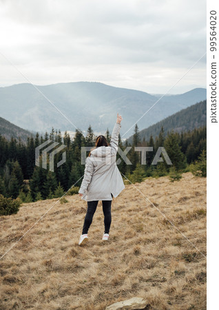 Happy girl stands in the mountains, enjoys the beautiful view and raises her hand up Happy girl stands in the mountains, enjoys the beautiful view and raises her hand up 99564020
