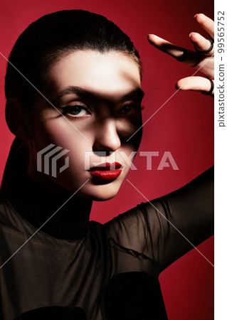 Beautiful woman in black clothes, classic make-up and red lips posing in the studio on a red background. 99565752