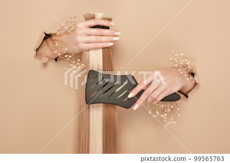 Hands with flowers hold strands of hair for extensions on a beige background. 99565763