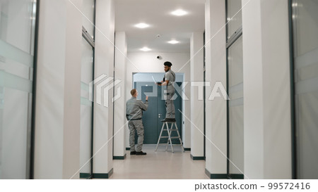 Two multi ethnic installers in uniform sets up security camera 99572416