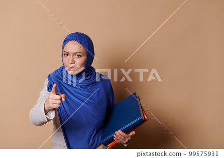 Confident strict Arab Muslim woman in blue hijab, pointing at camera, warning about the importance of reading book, standing with pile of books and looking at camera, isolated against beige background Confident strict Arab Muslim woman in blue hijab, pointing at camera, warning about the importance of reading book, standing with pile of books and looking at camera, isolated against beige background 99575931