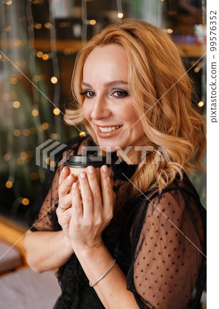 Young charming blonde with a cute smile and makeup while relaxing in a cafe. She is holding a cup of coffee in her hands. She is dressed in a black dress with transparent sleeves. Young charming blonde with a cute smile and makeup while relaxing in a cafe. She is holding a cup of coffee in her hands. She is dressed in a black dress with transparent sleeves. 99576352