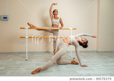 Near the barre, in the choreographic hall, two women in a beige top and leggings are engaged. Near the barre, in the choreographic hall, two women in a beige top and leggings are engaged. 99576379