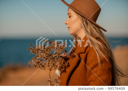 A woman walking along the coast near the sea. An elegant lady in a brown coat and a hat with fashionable makeup walks on the seashore 99576409