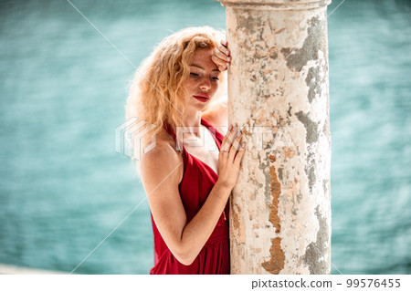 Outdoor portrait of a young beautiful natural redhead girl with freckles, long curly hair, in a red dress, posing against the background of the sea. 99576455