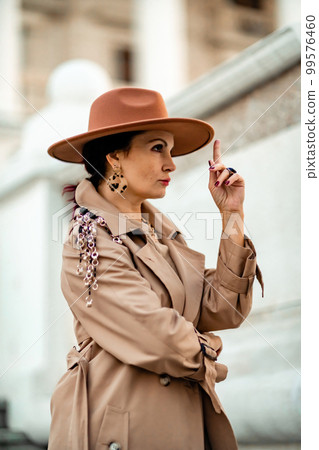 Outdoor fashion portrait of young elegant fashionable brunette woman, model in stylish hat, choker and light raincoat posing at sunset in European city. Outdoor fashion portrait of young elegant fashionable brunette woman, model in stylish hat, choker and light raincoat posing at sunset in European city. 99576460
