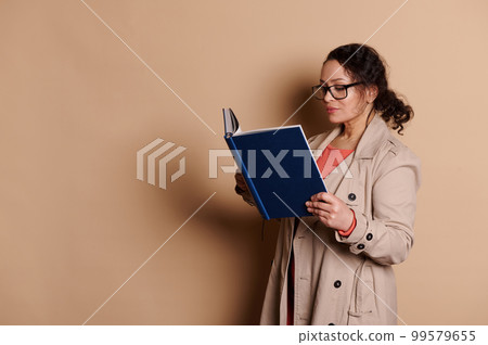Middle-aged multi-ethnic brunette woman wearing elegant beige coat and stylish eyeglasses, reading a book, on isolated beige background. Education. Learning. Erudition and people concept. Copy space 99579655