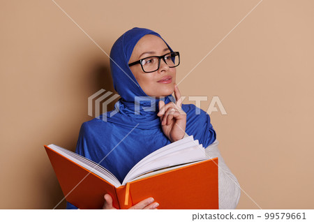 Beautiful Middle-Eastern Muslim woman wearing a blue hijab and eyeglasses, dreamily and thoughtfully looking aside, posing with a hardcover orange book, on isolated beige background. World Book Day Beautiful Middle-Eastern Muslim woman wearing a blue hijab and eyeglasses, dreamily and thoughtfully looking aside, posing with a hardcover orange book, on isolated beige background. World Book Day 99579661