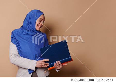Confident Muslim woman with head covered in hijab, smiling while holding pile of hardcover books in her hands, isolated on beige background with copy space. Portrait of a teacher, professor, educator 99579673