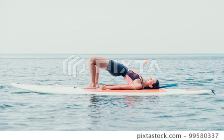 Woman sup yoga. Happy young sporty woman practising yoga pilates on paddle sup surfboard. Female stretching doing workout on sea water. Modern individual female outdoor summer sport activity. Woman sup yoga. Happy young sporty woman practising yoga pilates on paddle sup surfboard. Female stretching doing workout on sea water. Modern individual female outdoor summer sport activity. 99580337