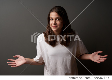 Smiling confused teen girl shrugs her shoulders with hands spreaded. 99582183