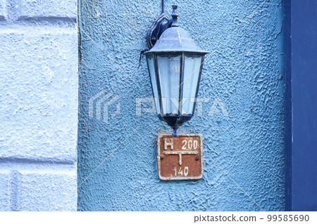 Old-fashioned street wall lamp made of black metal on blue wall facade of an old building with column and informational plaque Old-fashioned street wall lamp made of black metal on blue wall facade of an old building with column and informational plaque 99585690