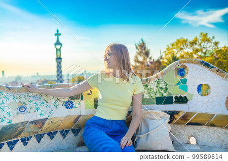 Positive young female tourist sitting on stone bench and checking photos while resting during vacation at Park Guell Positive young female tourist sitting on stone bench and checking photos while resting during vacation at Park Guell 99589814