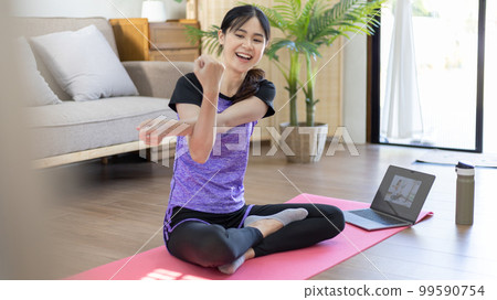 Woman looking at yoga teacher on laptop and doing light exercise in the living room at home, Watch live teaching or VDO on laptop, Dumbbells and a protein shake or bottle of water, Healthy exercise. 99590754