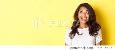 Image of excited and hopeful african-american girl in white t-shirt, smiling and looking amazed at upper left corner advertisement, standing over yellow background Image of excited and hopeful african-american girl in white t-shirt, smiling and looking amazed at upper left corner advertisement, standing over yellow background 99593687