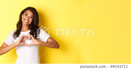 Image of lovely african-american girl in white t-shirt, showing heart sign and smiling, standing over yellow background Image of lovely african-american girl in white t-shirt, showing heart sign and smiling, standing over yellow background 99593711