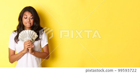 Portrait of attractive african-american woman, looking tempted at money, wanting to buy something, standing over yellow background 99593722