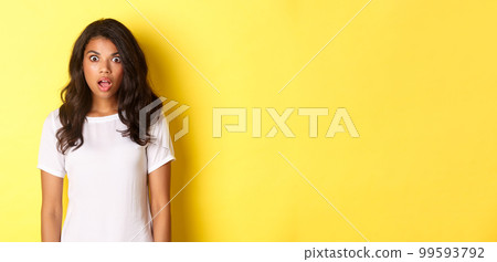 Portrait of shocked and speechless african-american girl, drop jaw and looking at amazing promo offer, standing over yellow background with copy space 99593792