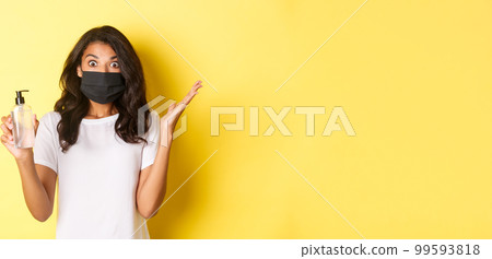 Concept of covid-19, social distancing and lifestyle. Image of excited african-american woman, wearing face mask, raising hands up surprised, holding hand sanitizer, yellow background 99593818