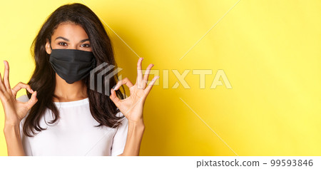 Concept of coronavirus, pandemic and lifestyle. Close-up of confident, smiling african-american woman in black face mask, showing okay signs in approval, like something good, yellow background 99593846