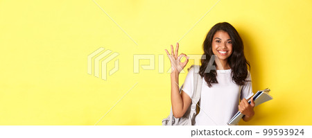 Portrait of satisfied african-american female student, smiling pleased and showing okay sign, like something good, standing over yellow background 99593924