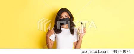 Concept of covid-19, social distancing and lifestyle. Portrait of cheerful african-american girl in face mask, recommend to use antiseptic, showing hand sanitizer and thumbs-up Concept of covid-19, social distancing and lifestyle. Portrait of cheerful african-american girl in face mask, recommend to use antiseptic, showing hand sanitizer and thumbs-up 99593925
