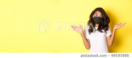 Concept of coronavirus, pandemic and lifestyle. Image of cute african-american girl in face mask, shrugging and looking clueless, dont know anything, standing over yellow background 99593998