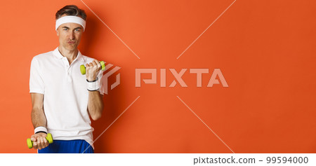 Portrait of cheeky middle-aged guy training in gym, lifting dummbells and smiling, workout against orange background 99594000