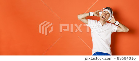 Portrait of carefree middle-aged male athlete, listening music in headphones during fitness training, standing over orange background Portrait of carefree middle-aged male athlete, listening music in headphones during fitness training, standing over orange background 99594010