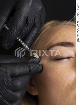 Closeup of Caucasian woman gets botox cosmetic injection in eye area. Beauty physician inserts needle of syringe into face of beautiful female. Cosmetologist, aesthetic medicine. Vertical Closeup of Caucasian woman gets botox cosmetic injection in eye area. Beauty physician inserts needle of syringe into face of beautiful female. Cosmetologist, aesthetic medicine. Vertical 99594520