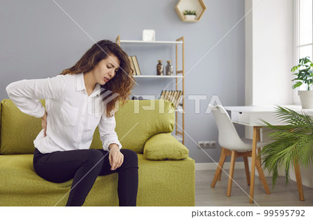 Woman with lumbago back pain sitting on the sofa and holding her hand on her lower back Woman with lumbago back pain sitting on the sofa and holding her hand on her lower back 99595792