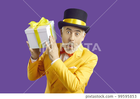 Senior man in yellow suit and hat with funny surprised face expression holding gift box 99595853
