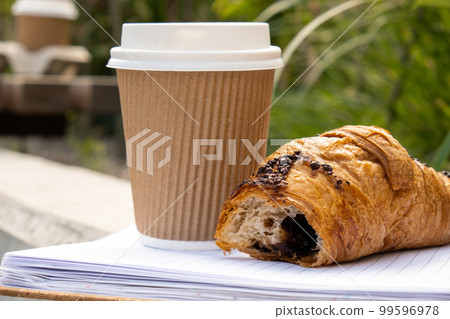 Fresh baked chocolate croissant on paper cup with lid for tea to go. Empty paper notebook. Freelancer's place of work. Study and work. Remote business education. Making plans for next week year. Being 99596978